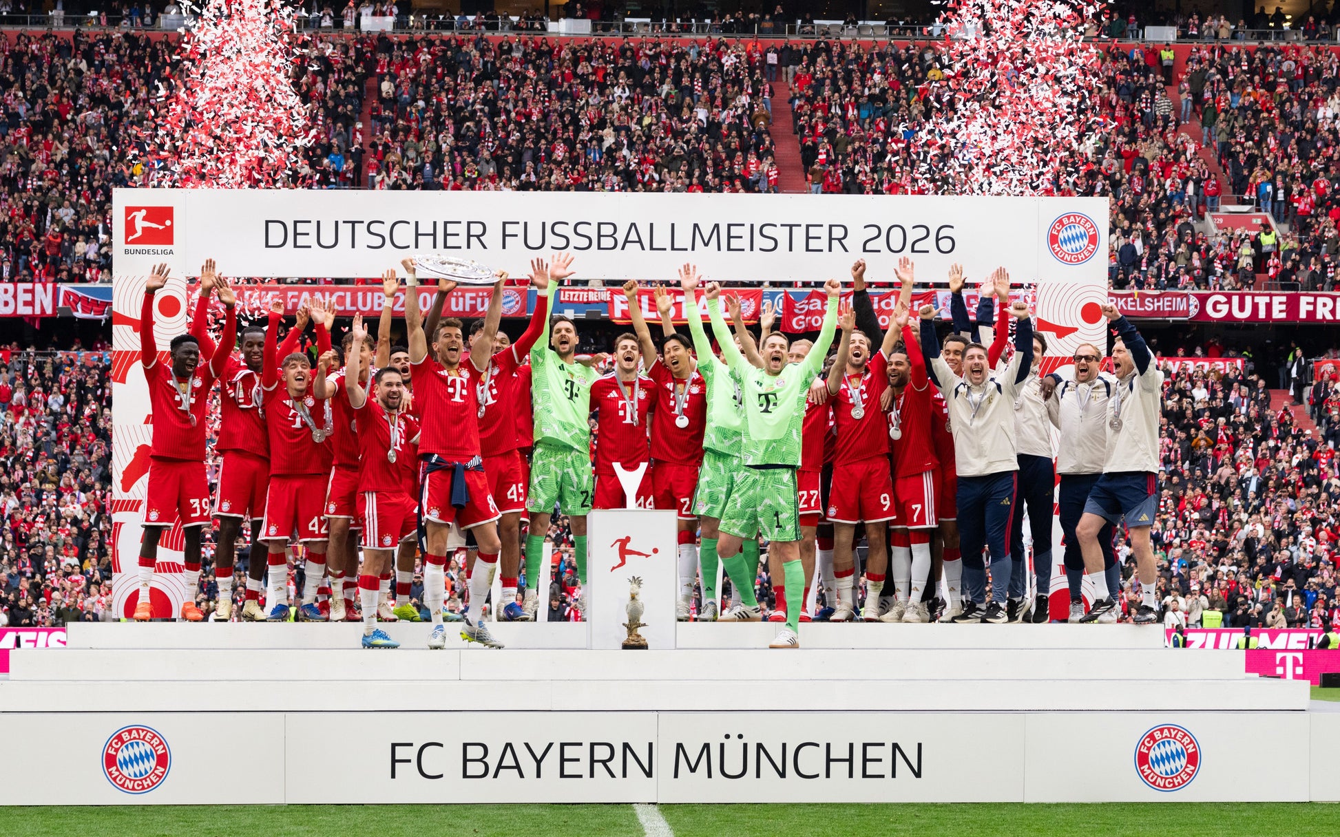 FC Bayern Munich football team celebrating on a podium with confetti and fireworks in a stadium. Players in red jerseys and a goalkeeper in green stand behind a banner reading 'DEUTSCHER FUSSBALLMEISTER 2026'.