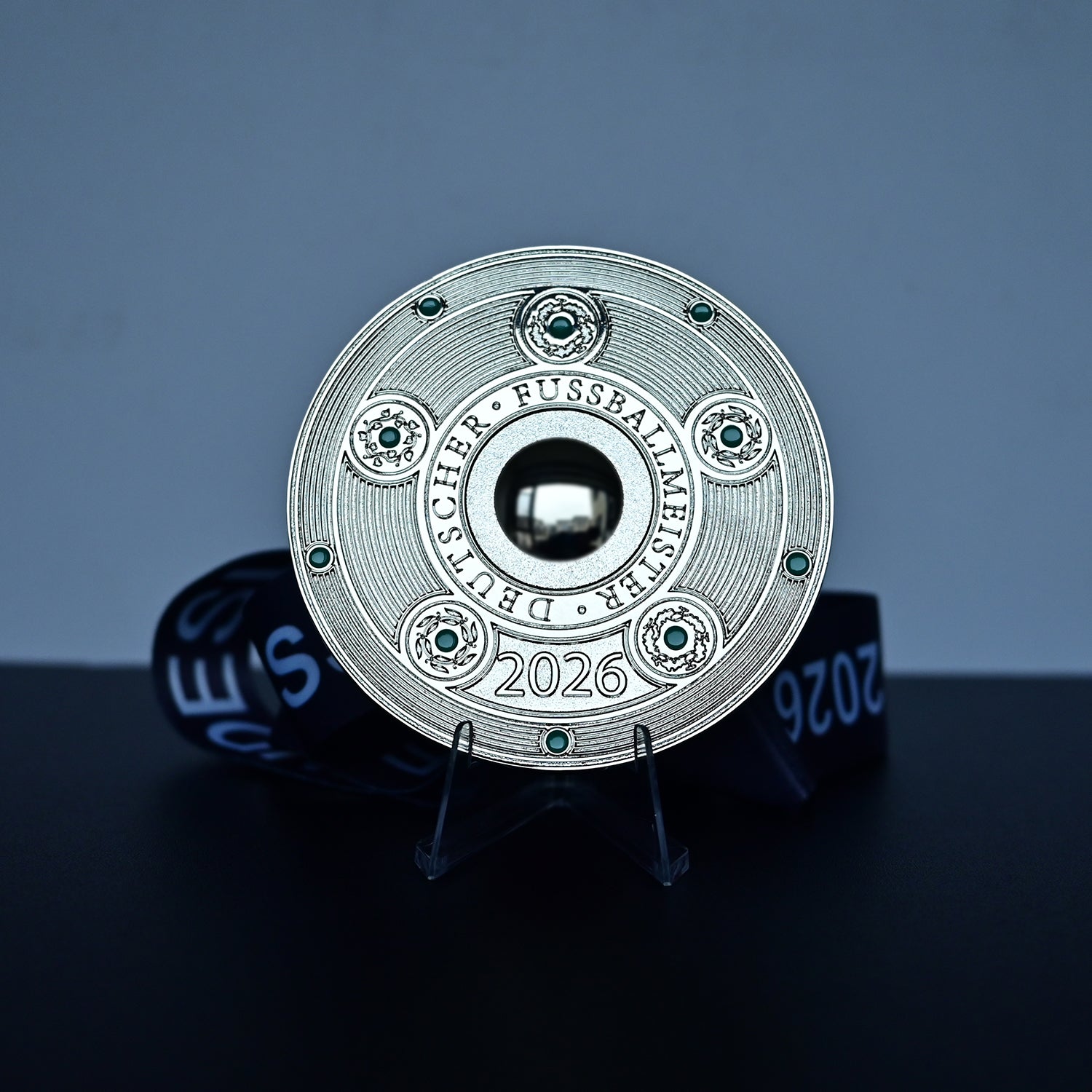 A round silver-toned football award plaque for the 2026 FIFA World Cup in Germany, displayed on a clear stand with a dark blue ribbon behind it against a blue-gray background.