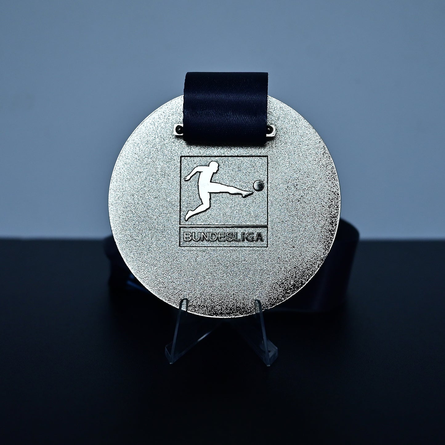 A round silver medal with a black ribbon, displayed on a clear stand against a dark gray background. The medal features an embossed soccer player logo with the text 'BUNDESliga' underneath.