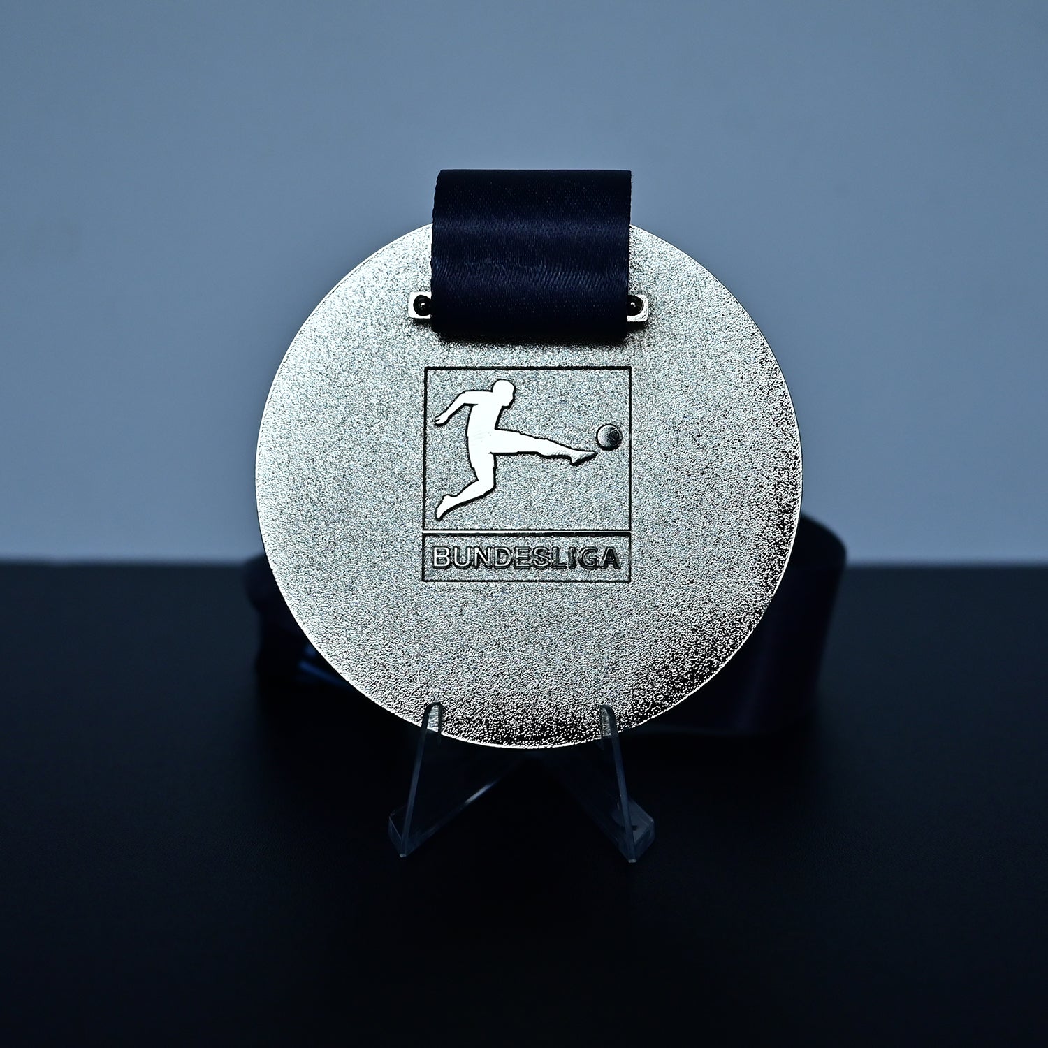 A round silver medal with a black ribbon, displayed on a clear stand against a dark gray background. The medal features an embossed soccer player logo with the text 'BUNDESliga' underneath.