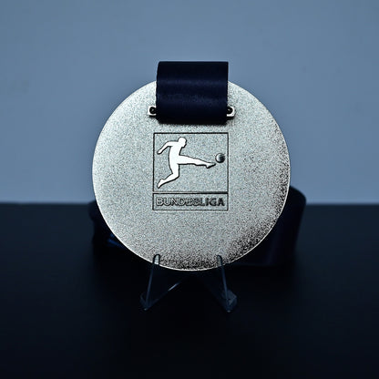 A round silver medal with a black ribbon, displayed on a clear stand against a dark gray background. The medal features an embossed soccer player logo with the text 'BUNDESliga' underneath.