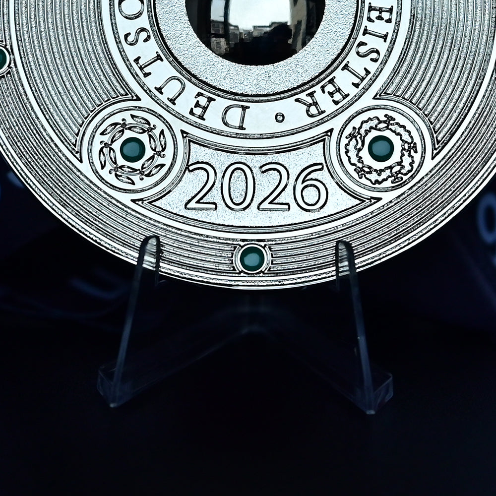 A close-up of a round silver-toned commemorative coin or medallion displayed on a clear stand against a dark background. The coin features the text 'EISTER • DEUTSCHLAND' around the top edge, '2026' in the center, and decorative circular details with small green accents.