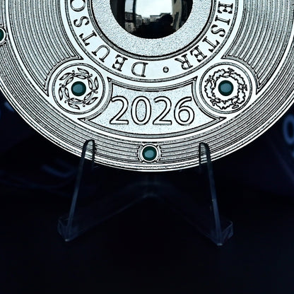 A close-up of a round silver-toned commemorative coin or medallion displayed on a clear stand against a dark background. The coin features the text 'EISTER • DEUTSCHLAND' around the top edge, '2026' in the center, and decorative circular details with small green accents.