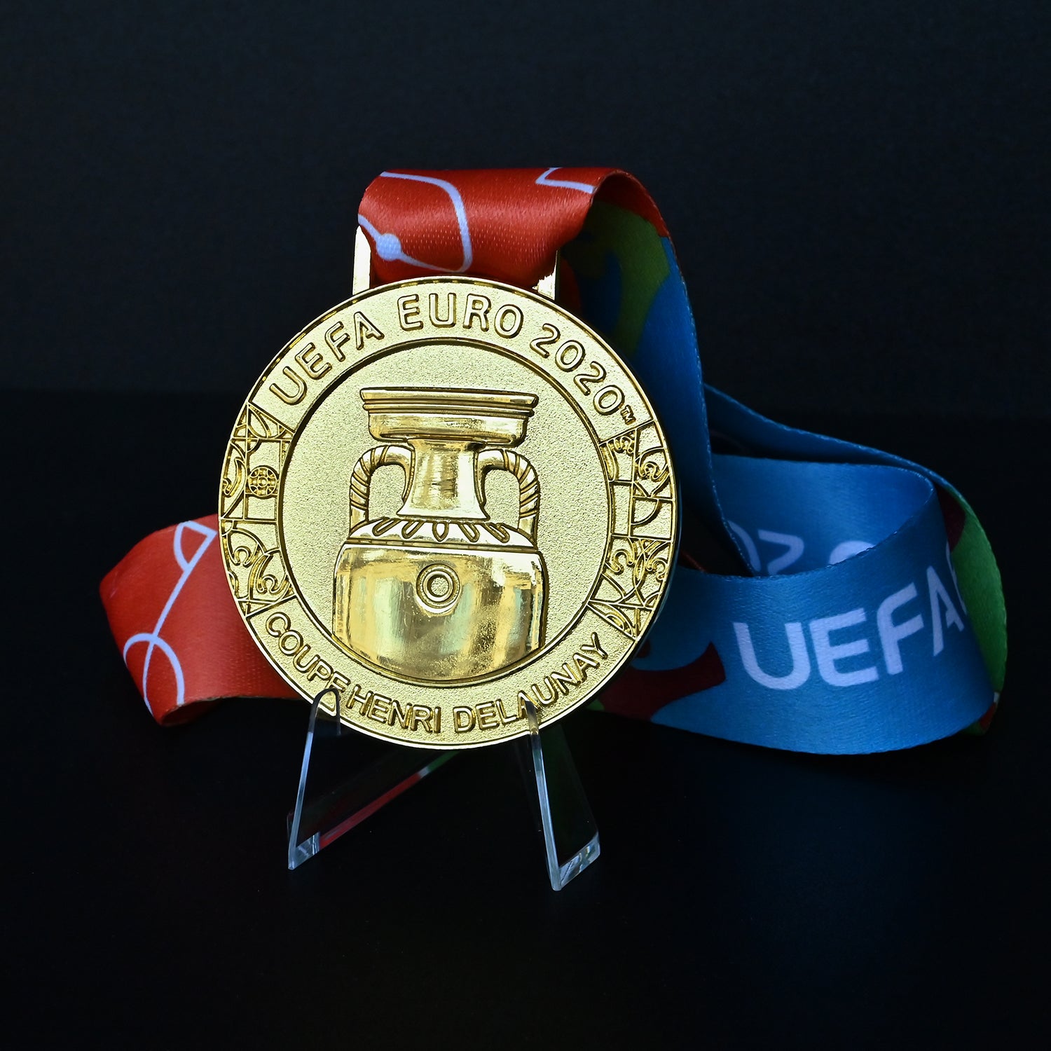 UEFA Euro 2020 championship medal with iconic Coupe Henri Delaunay engraving, red-blue-green tournament ribbon, displayed on clear acrylic stand. Premium Italy victory commemorative collectible.